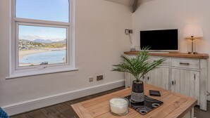 Una habitación con un amplio ventanal arqueado con vista a la playa, un televisor sobre un mueble de madera y una mesa con una vela y una planta.