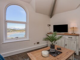 Una habitación con un amplio ventanal arqueado con vista a la playa, un televisor sobre un mueble de madera y una mesa con una vela y una planta.