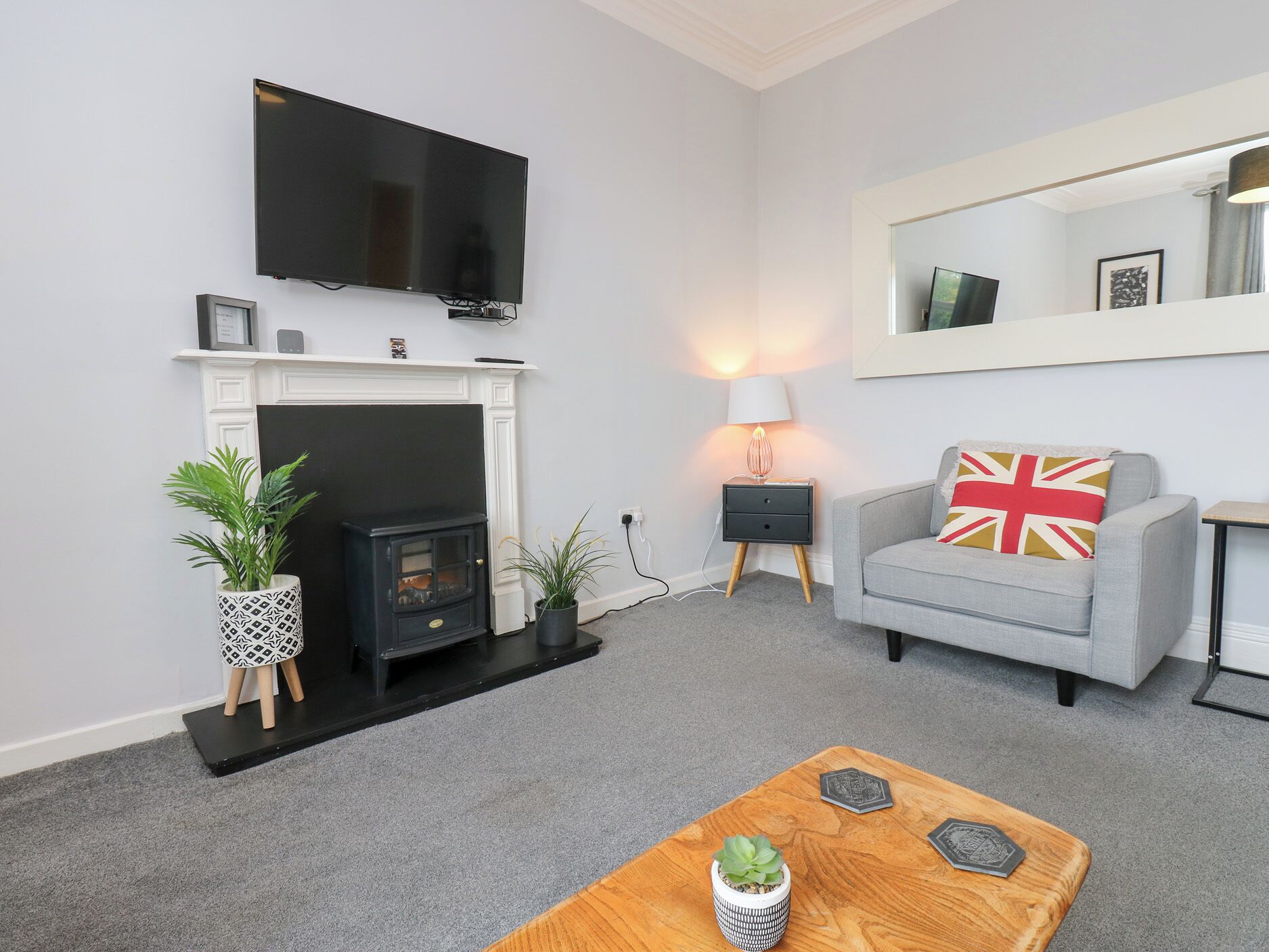 Un salón con chimenea, un televisor montado encima, un sofá gris con un cojín de la bandera del Reino Unido y una mesa de centro de madera con objetos decorativos.