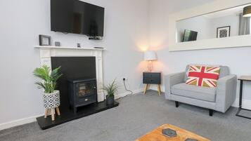 Un salón con chimenea, un televisor montado encima, un sofá gris con un cojín de la bandera del Reino Unido y una mesa de centro de madera con objetos decorativos.