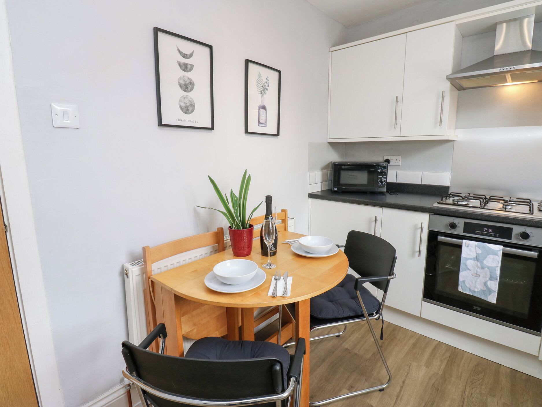 A compact kitchen with white cabinets, a microwave, an oven, and a dining table with chairs. There are framed pictures on the wall above the table.