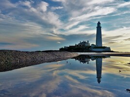 Un faro con reflejo claro en aguas tranquilas, una costa rocosa y un edificio con tejados rojos al fondo.