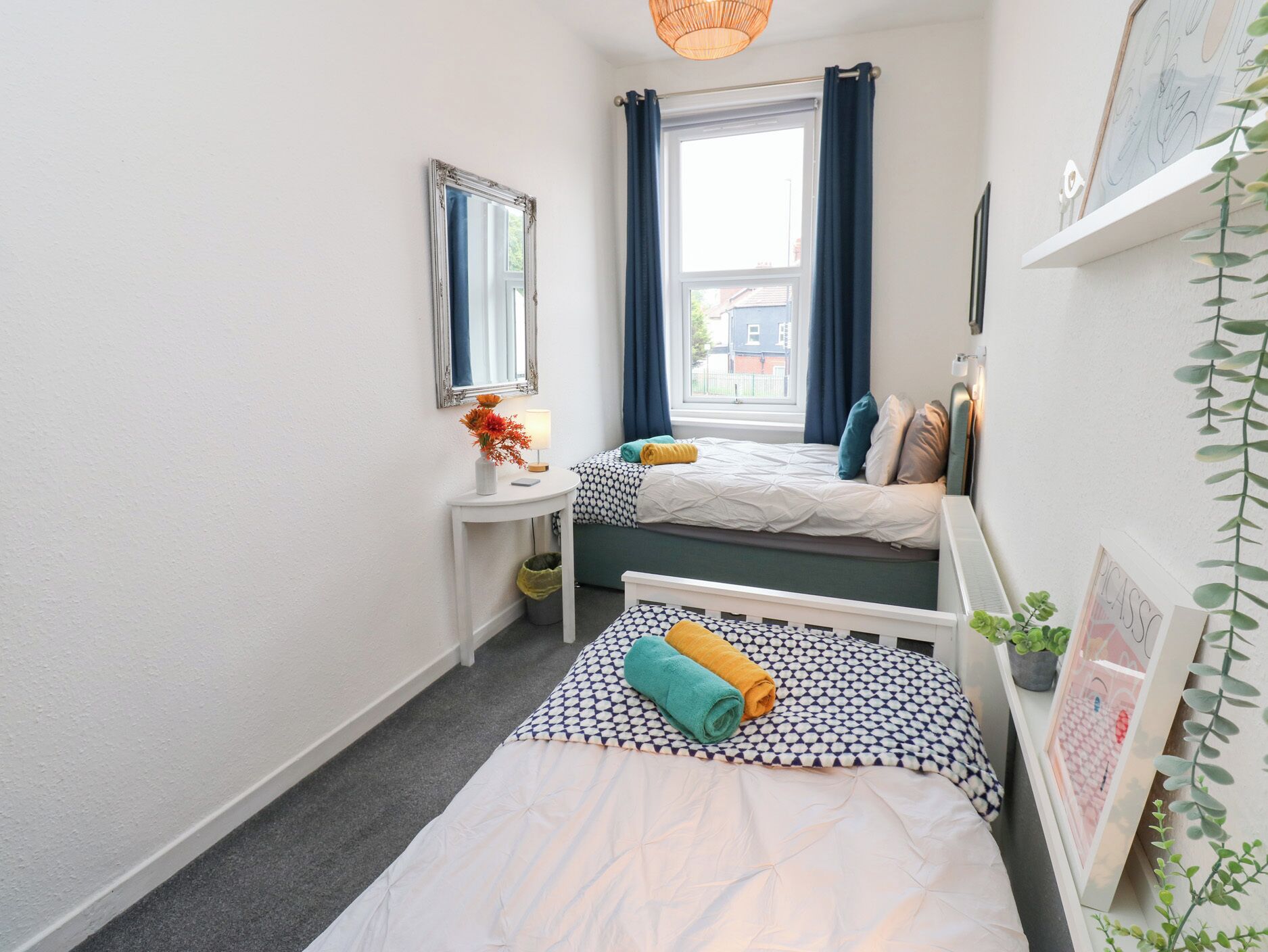 A small, well-lit room with two beds, a bedside table, a mirror, and a window with curtains.