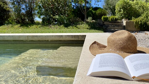 Une piscine aux eaux limpides, bordée de pierres, un chapeau de paille et un livre ouvert posé sur le bord.