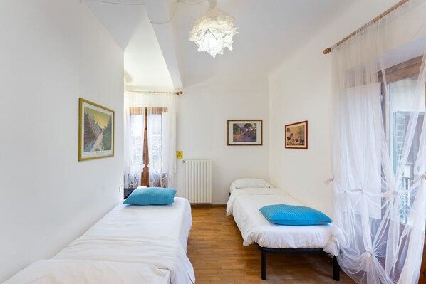Una habitación con dos camas individuales, un radiador y una ventana con cortinas traslúcidas.