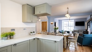 Eine moderne Küche mit grauen Schränken, einer weißen Arbeitsplatte und Holzböden. Es gibt einen Essbereich mit Tisch und Stühlen sowie ein Wohnzimmer mit einem blauen Sofa und einem an der Wand befestigten Fernseher.