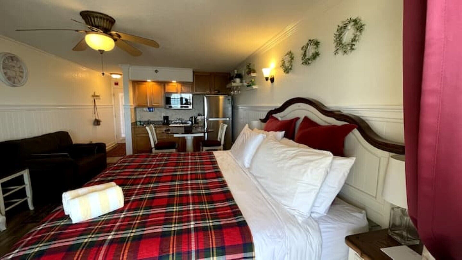 Une chambre à coucher comprenant un lit, une table de chevet, une chaise, une petite table, un ventilateur de plafond, un coin cuisine et une horloge murale.