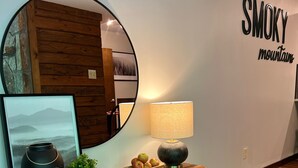Ein modernes Interieur mit einem Holzkonsolentisch, einem runden Spiegel, einer Wandleuchte, einem gerahmten Bild, einer Obstschale und einer Topfpflanze.