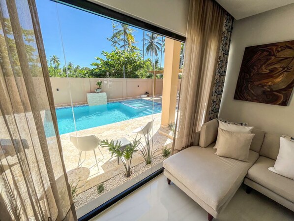 A modern living room with a large sliding glass door leading to a private pool area, a beige sofa, and a painting on the wall.