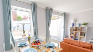 A dining area with a wooden table set for two, a bowl of fruit, and a bowl of pastries. There is a white chair and an orange sofa in the background. - Mermaid House (Weymouth)