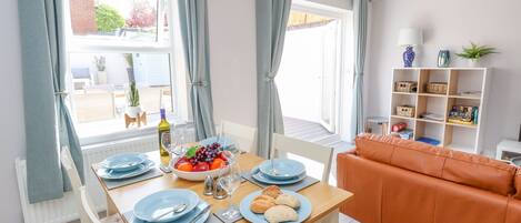 A dining area with a wooden table set for two, a bowl of fruit, and a bowl of pastries. There is a white chair and an orange sofa in the background.