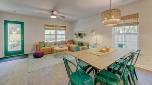 A spacious living room with a large wooden dining table, green chairs, a sofa with cushions, a ceiling fan, and large windows.