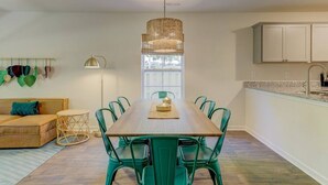 A dining area with a wooden table and green chairs, a hanging light fixture, and a window with blinds.