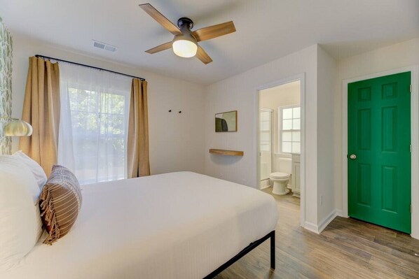Une chambre à coucher avec un lit, un ventilateur de plafond, une fenêtre avec des rideaux, une porte verte et une salle de bain visible à travers une porte ouverte.