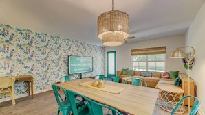 A room with a wooden dining table, teal chairs, a woven pendant light, a sofa with cushions, and a wall-mounted TV displaying "WELCOME TO SOUTH CAROLINA".
