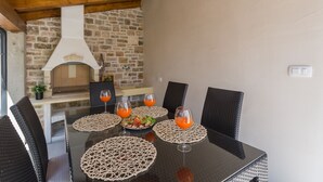 Ein Essbereich mit einer Steinwand, einem Backofen aus Ziegeln und Korbstühlen um einen Glastisch mit Untersetzern und Gläsern Orangensaft.