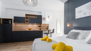 Ein modernes Hotelzimmer mit grauem Bett, Holzboden, Küchenzeile mit schwarzen Schränken und einer Holzarbeitsplatte sowie einer Wanduhr.