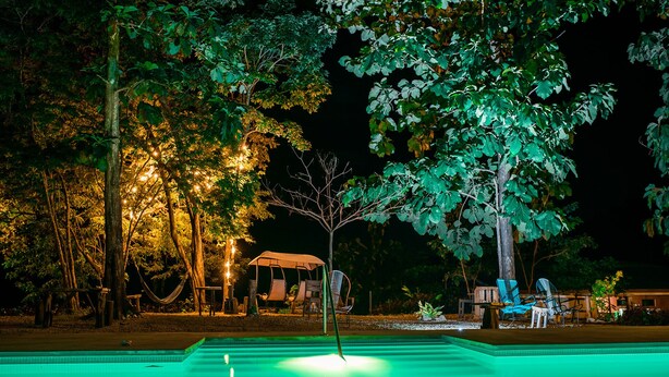 Un espace piscine aménagé pour la nuit, avec un arbre au centre, entouré de chaises et d’un hamac.