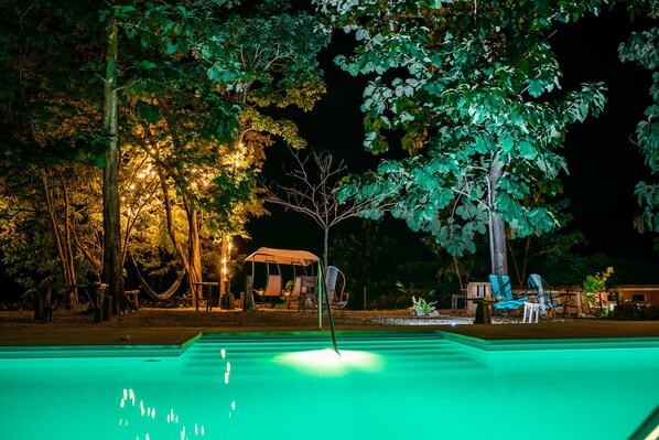 Ein Poolbereich bei Nacht mit einem Baum in der Mitte, umgeben von Stühlen und einer Hängematte.