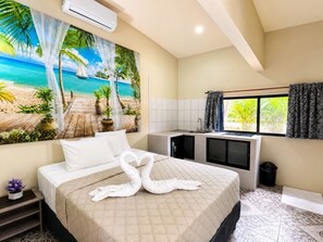 A hotel room with a bed, a painting of a beach scene, a kitchenette, and a bathroom.