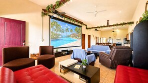 A modern living room with a large TV screen displaying a pool view, leather sofas, a coffee table, and a kitchen area in the background.