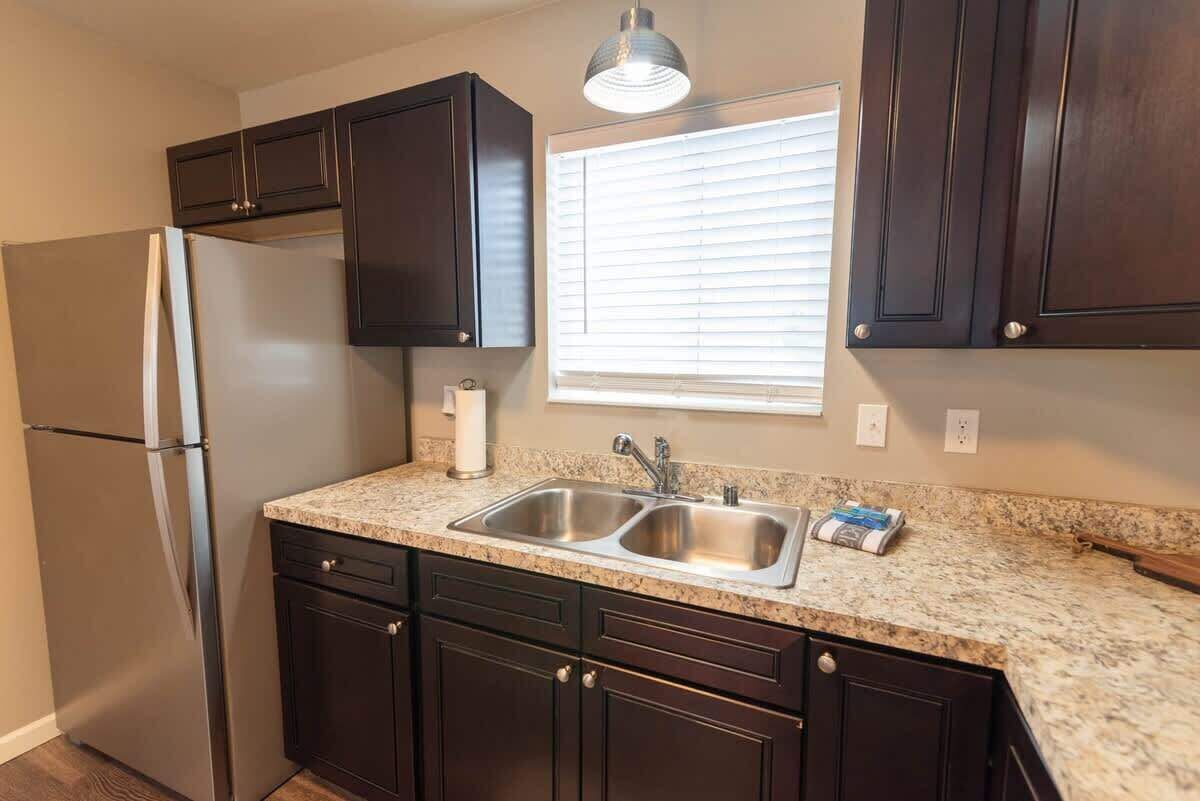 A kitchen with dark cabinets, a stainless steel refrigerator, a double sink, and a window with blinds.