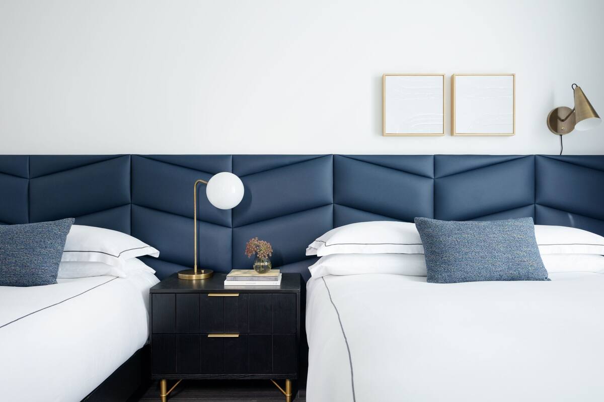 A modern bedroom with a dark blue headboard, two beds, a nightstand, a lamp, and two framed pictures on the wall.