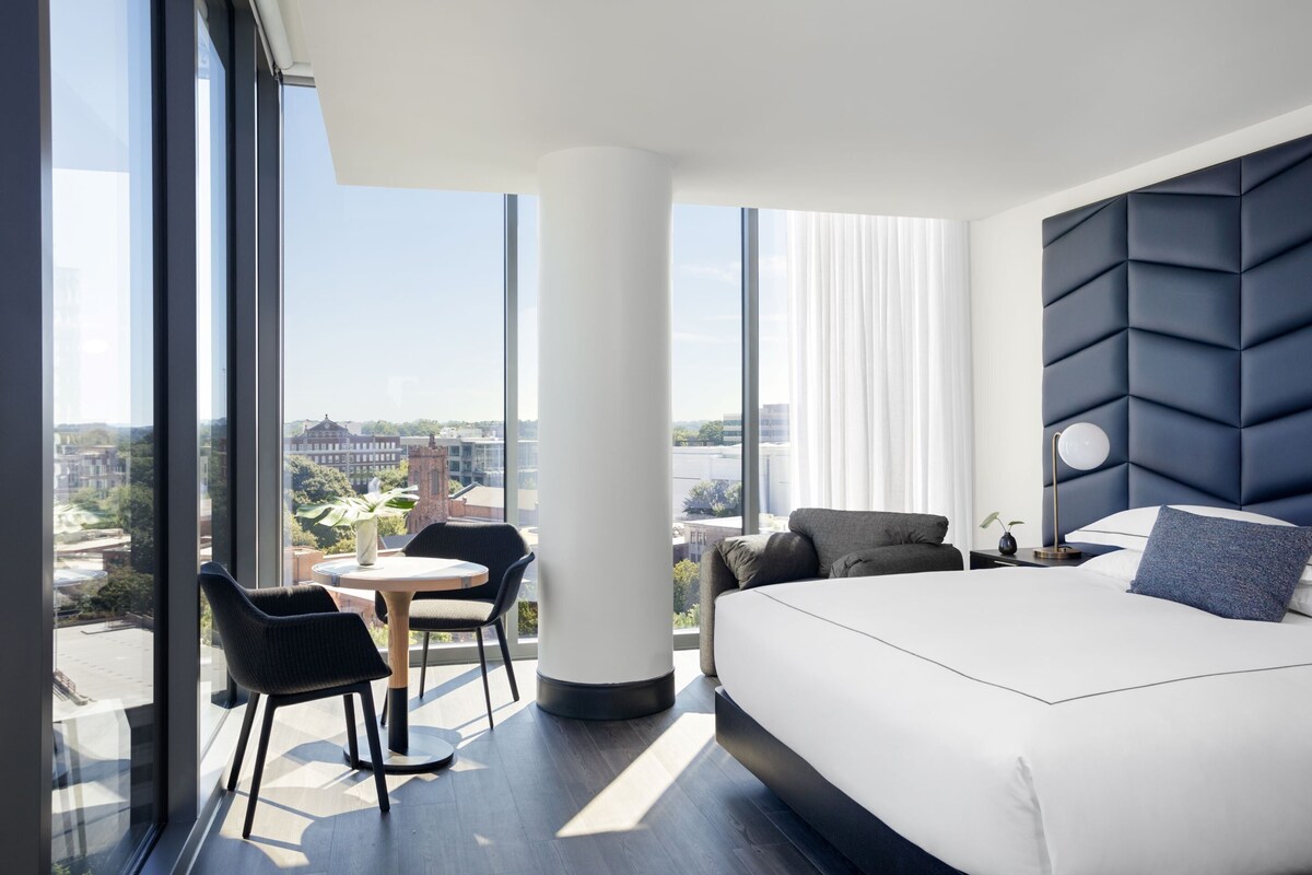 Habitación de hotel moderna con una cama grande, una mesa redonda y una silla. Un ventanal grande ofrece vistas de la ciudad.