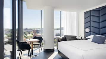 A modern hotel room with a large bed, a round table, and a chair. There is a large window offering a city view.