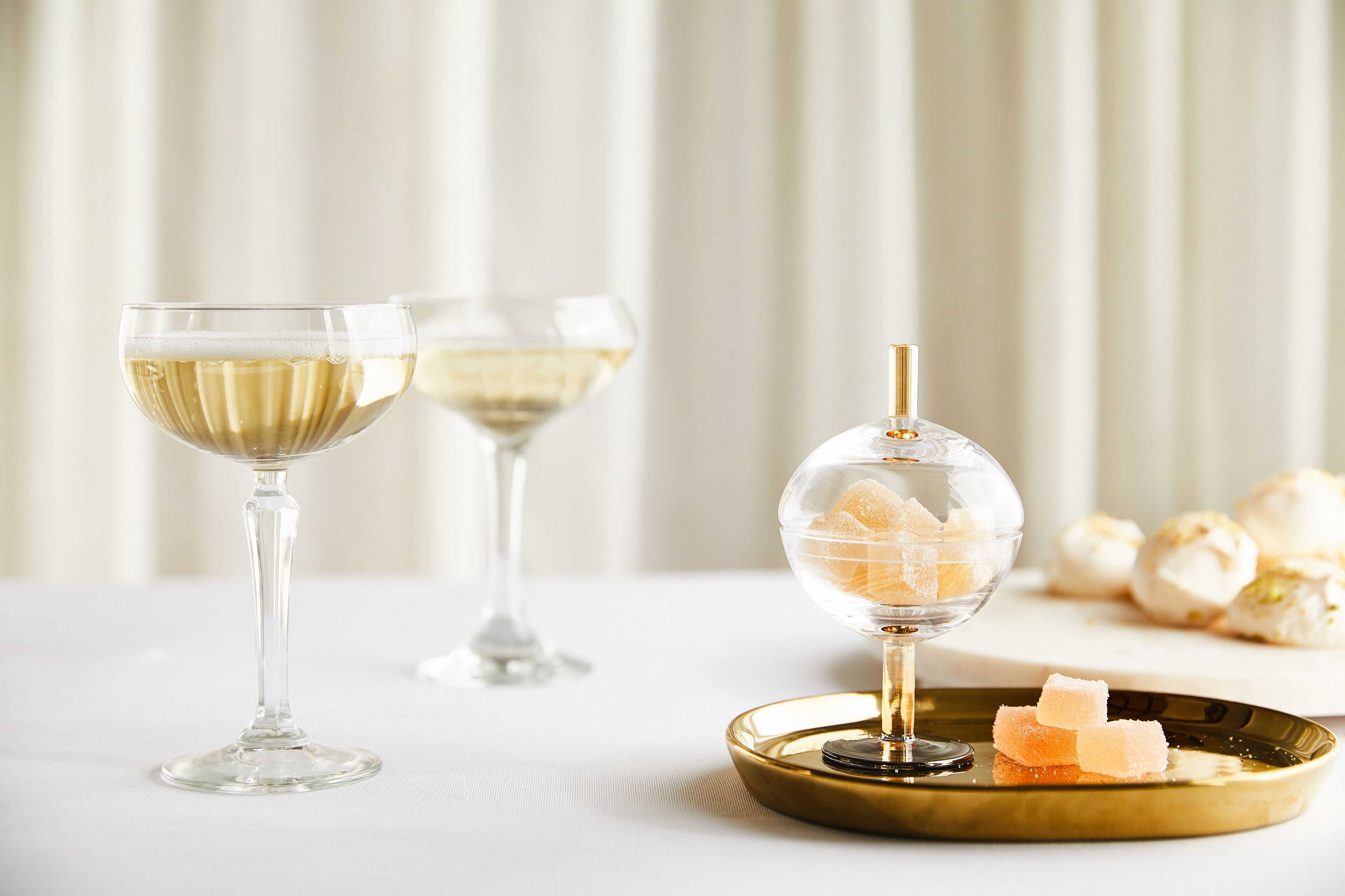 Two glasses of white wine on a table with a decorative glass containing sugar cubes and a plate with more sugar cubes.