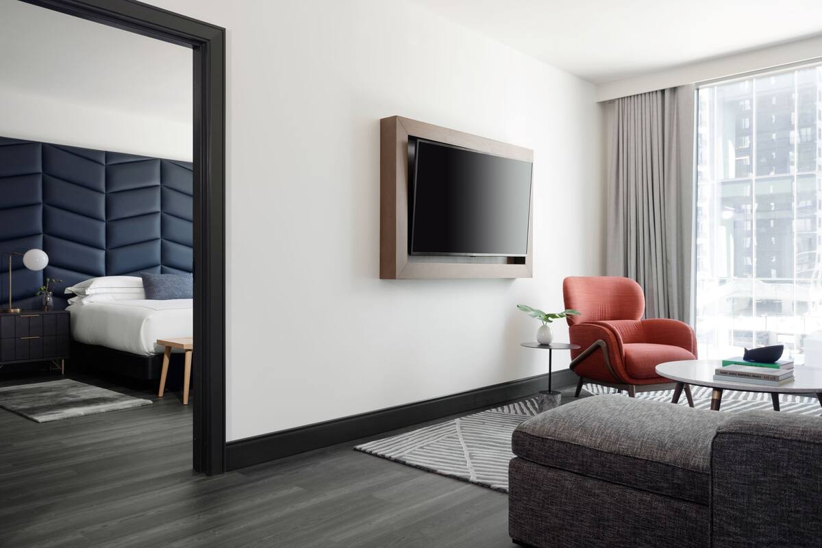 A modern hotel room with a bed, a TV mounted on the wall, a red armchair, a small table with books, and a large window with curtains.