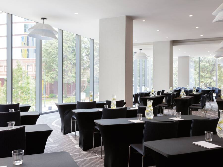 Une salle de conférence moderne, dotée de grandes fenêtres, de chaises et de tables noires, ainsi que de verres et d’un vase décoratif.