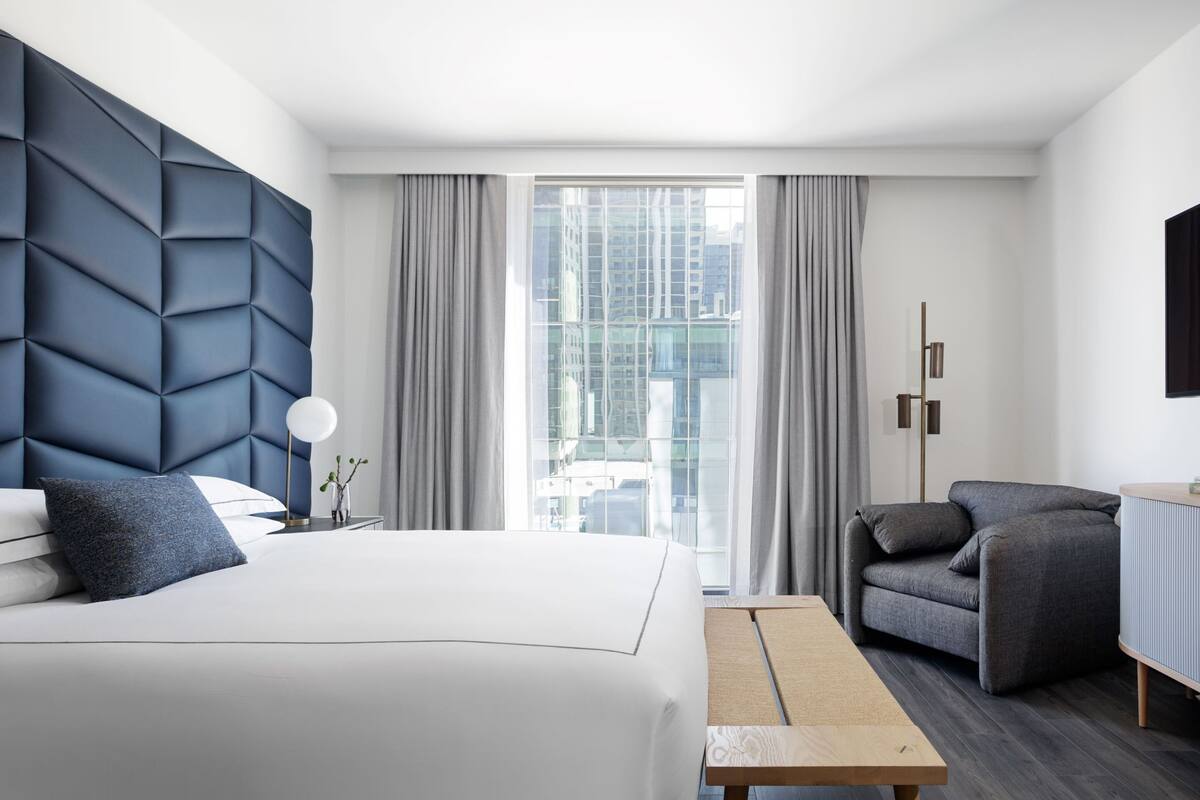 A modern hotel room with a large bed, a grey armchair, a small table, and a view of the city through large windows.