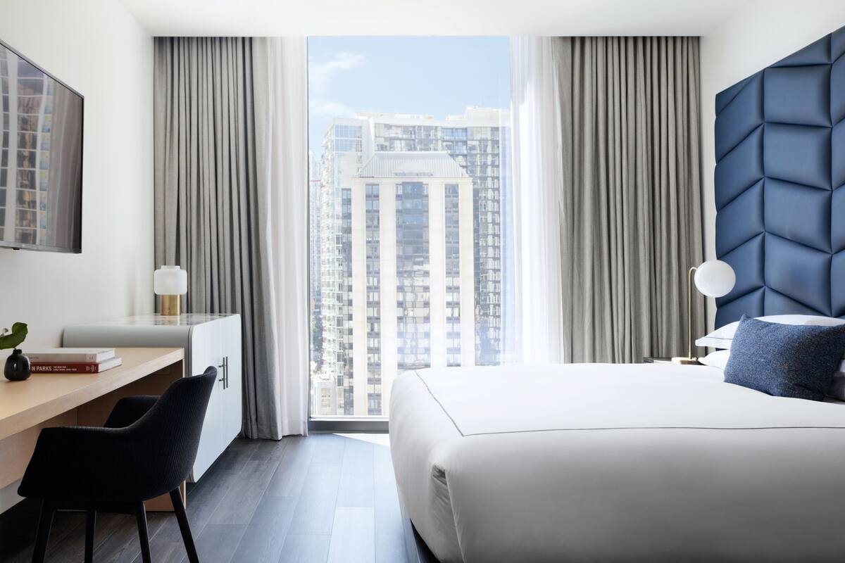 A modern hotel room with a large bed, a desk, and a view of a cityscape through floor-to-ceiling windows.