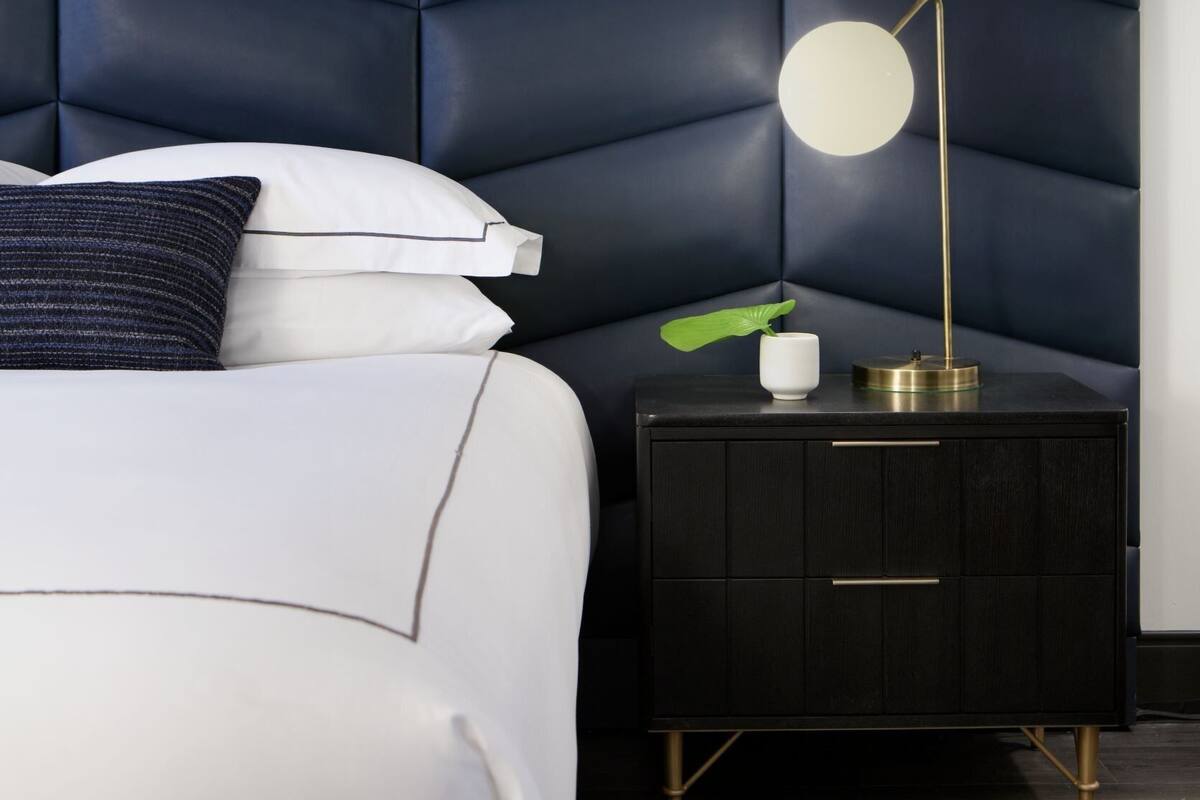 A bedroom with a dark blue padded headboard, a white bed with pillows, a bedside table with a lamp, and a small plant.