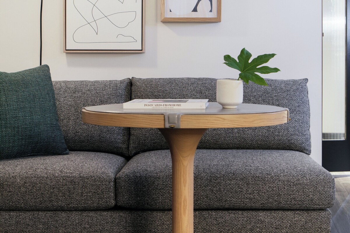 Una sala de estar moderna con un sofá gris, una mesa redonda de madera con un libro y una planta, y dos obras de arte abstracto enmarcadas en la pared.