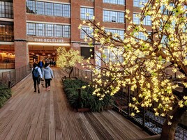 Un pasarela de madera con un edificio de ladrillo al fondo, un árbol con flores iluminadas y personas caminando.