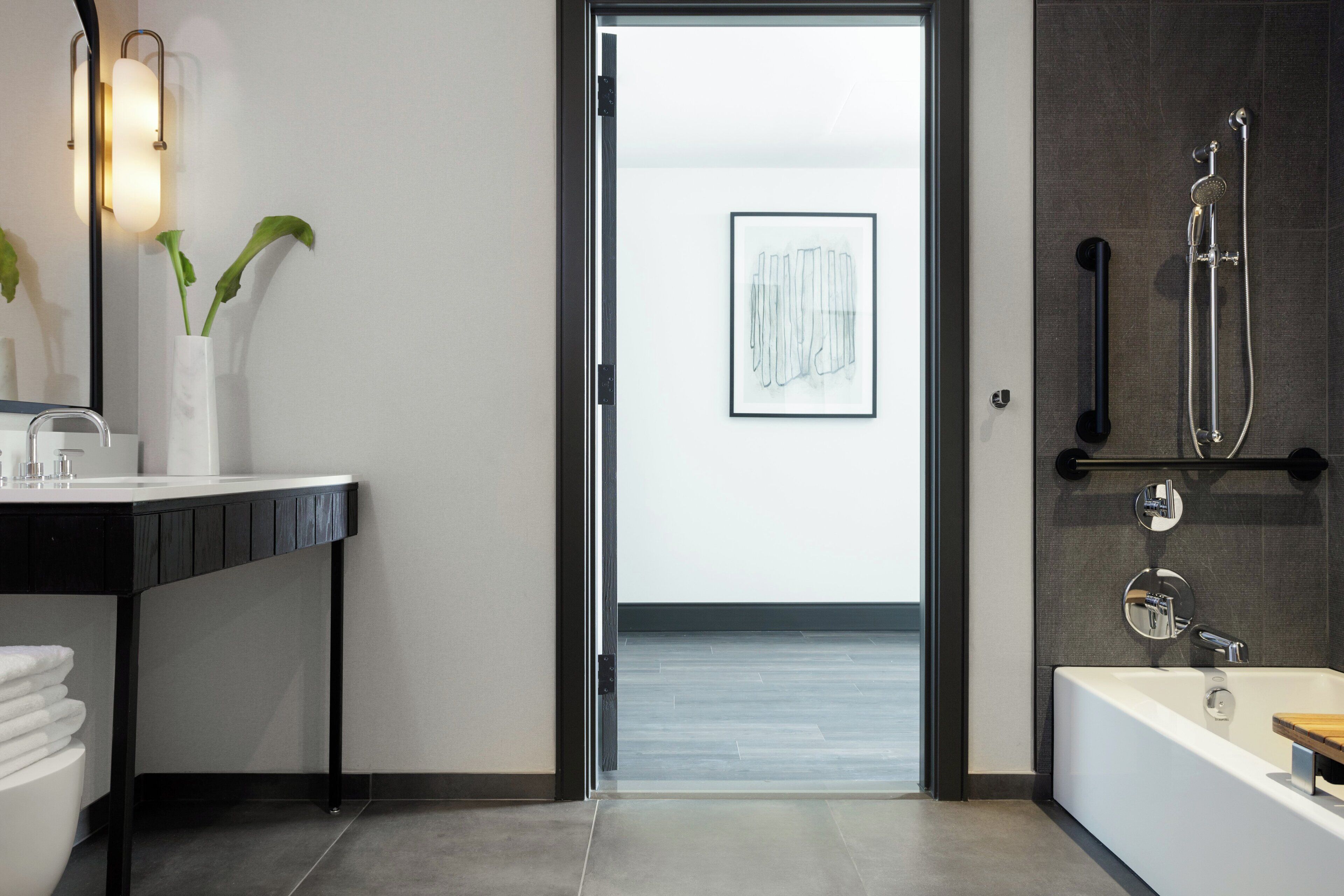 A modern bathroom with a black countertop, white sink, and a large mirror. There is a framed artwork on the wall and a shower area with a handheld showerhead.