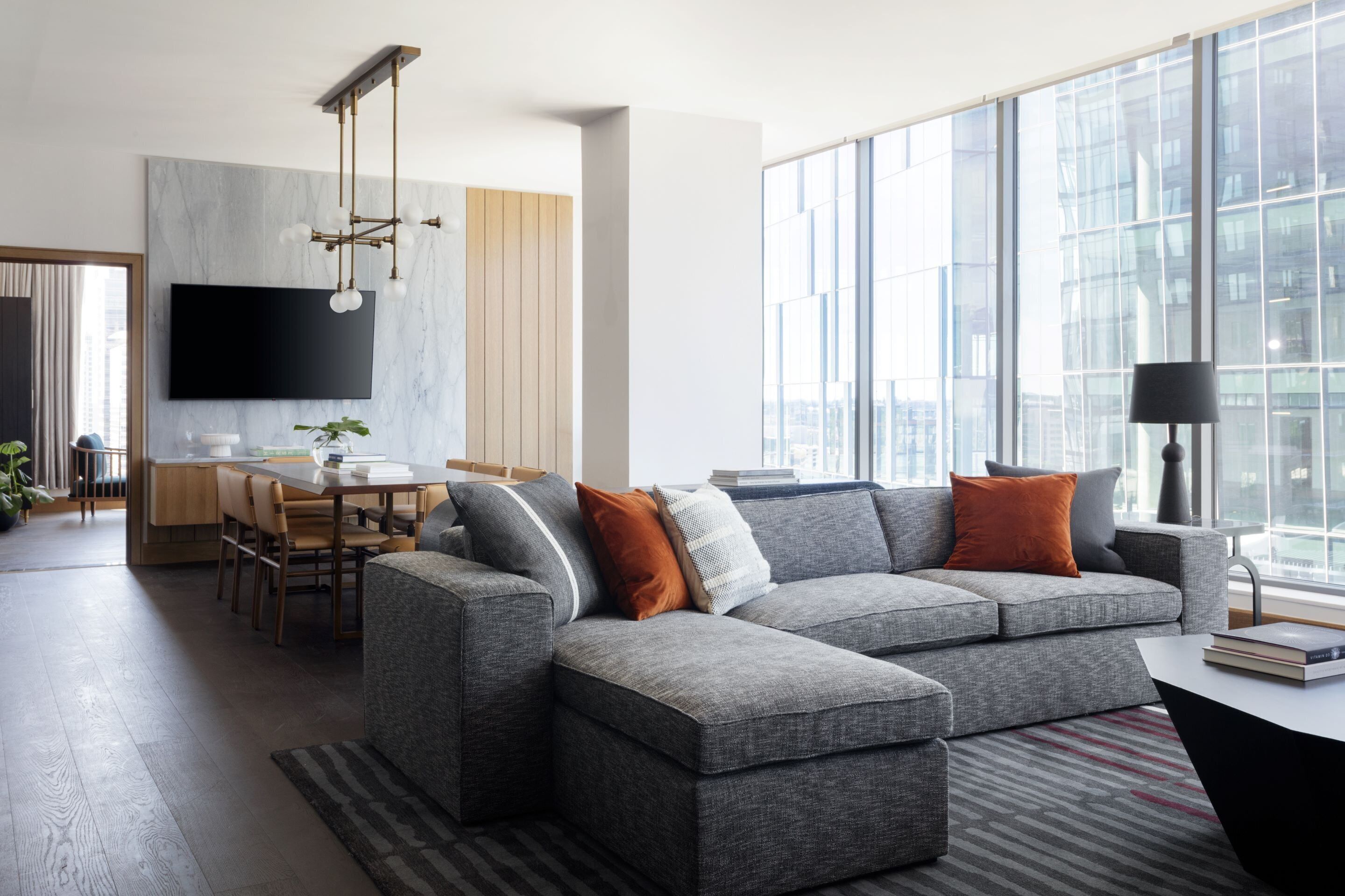 Un salon moderne avec un canapé d’angle gris, un coin repas avec une table et des chaises en bois, un téléviseur à écran plat fixé au mur et de grandes fenêtres offrant une vue sur la ville.