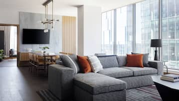 Un salon moderne avec un canapé d’angle gris, un coin repas avec une table et des chaises en bois, un téléviseur à écran plat fixé au mur et de grandes fenêtres offrant une vue sur la ville.