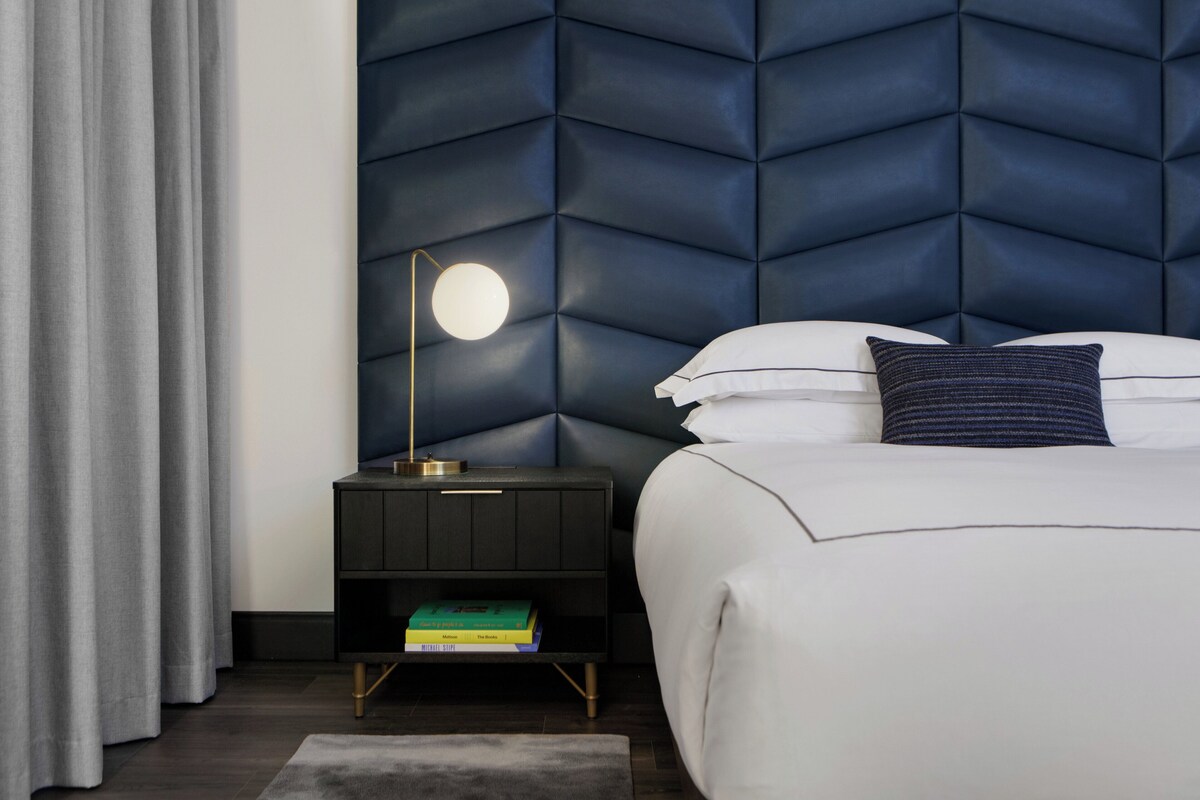 Un dormitorio moderno con cabecera capitonada, una mesita de noche con lámpara y una cama bien tendida con almohadas.