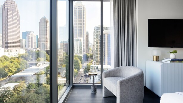 A modern hotel room with a large window offering a city view, a comfortable armchair, a flat-screen TV, and a small side table.