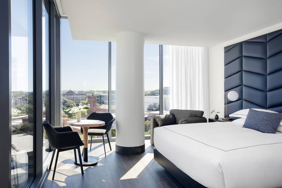 A modern hotel room with a large bed, a round table, and a chair. There is a large window offering a city view.