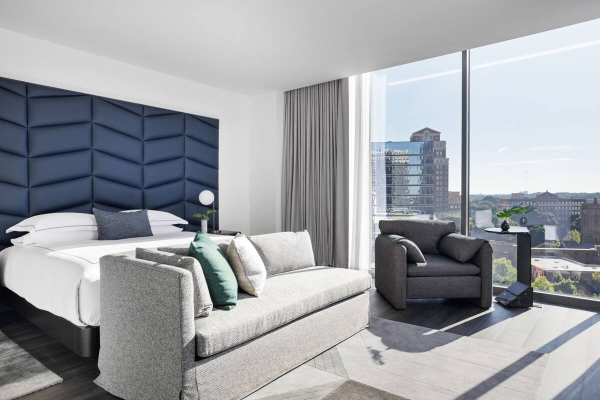 A modern hotel room with a large bed, a grey sofa, a grey armchair, and a large window offering a city view.
