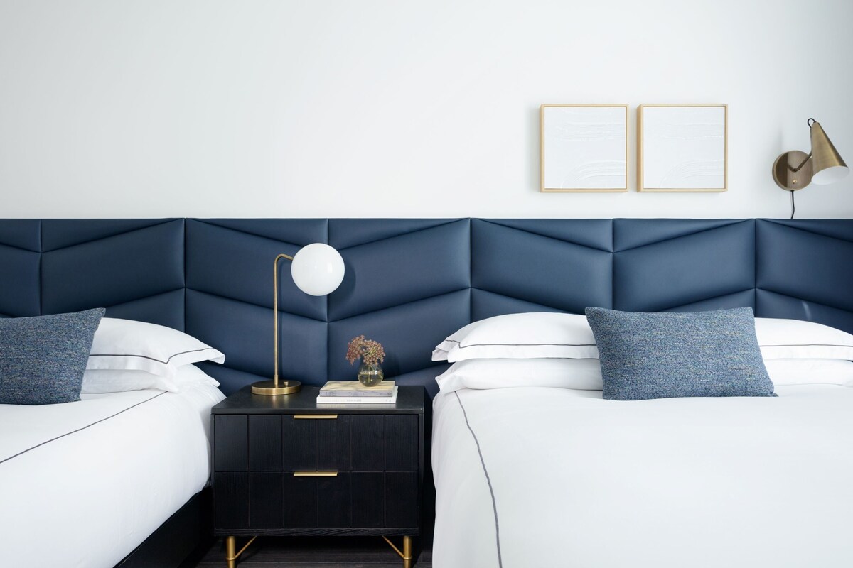Un dormitorio moderno con cabecera azul oscuro, dos camas, una mesita de noche, una lámpara y dos cuadros en la pared.
