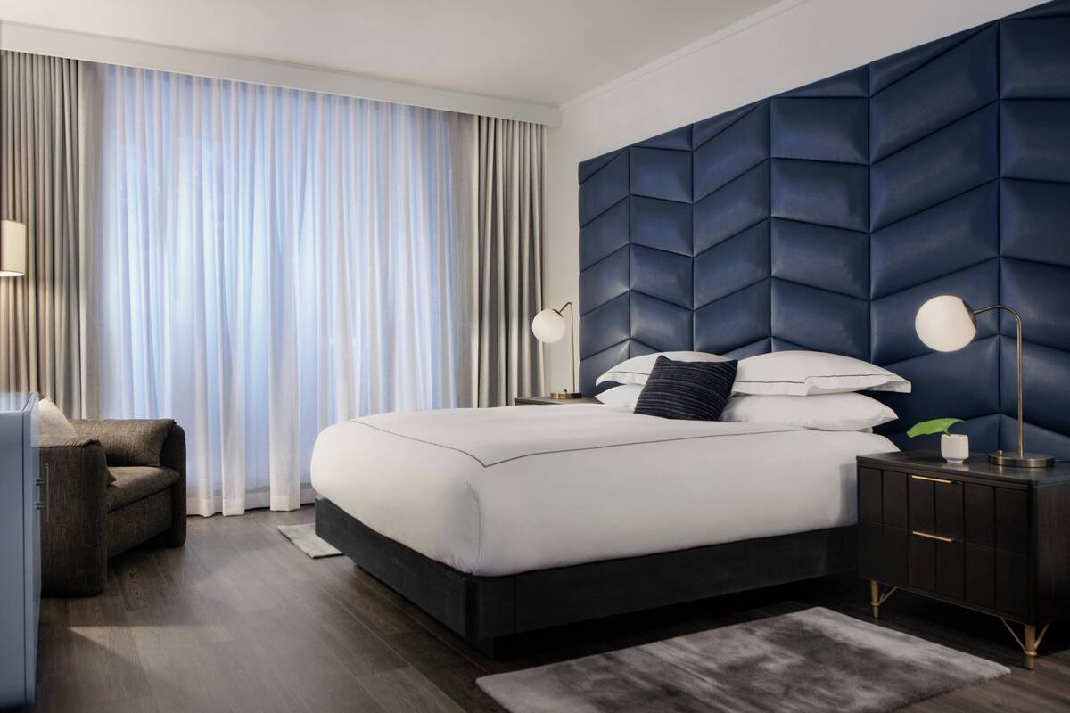A modern hotel room with a large bed, dark headboard, and a view of the city through floor-to-ceiling windows.