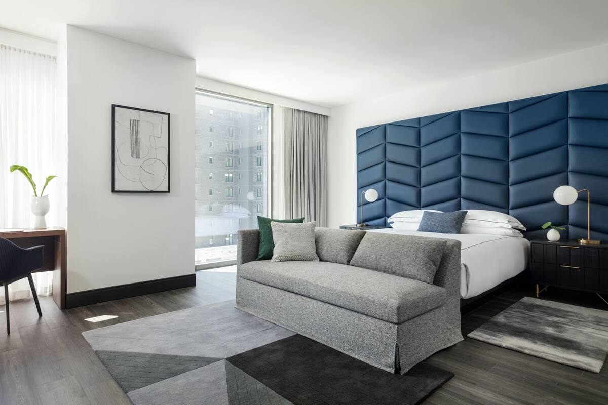 A modern bedroom with a grey sofa, a bed with a blue headboard, a bedside table with a lamp, and a large window with curtains.