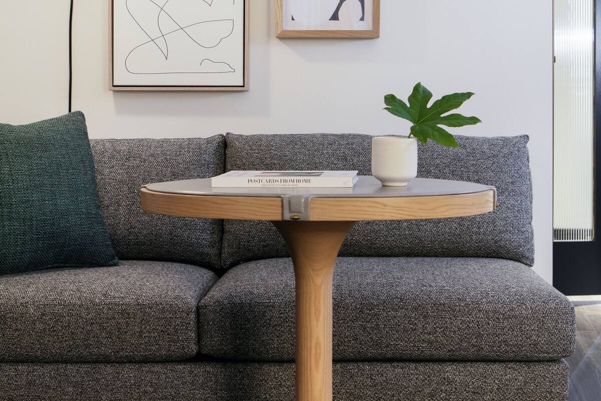 A modern living room with a grey sofa, a round wooden table with a book and a plant, and two framed abstract artworks on the wall.
