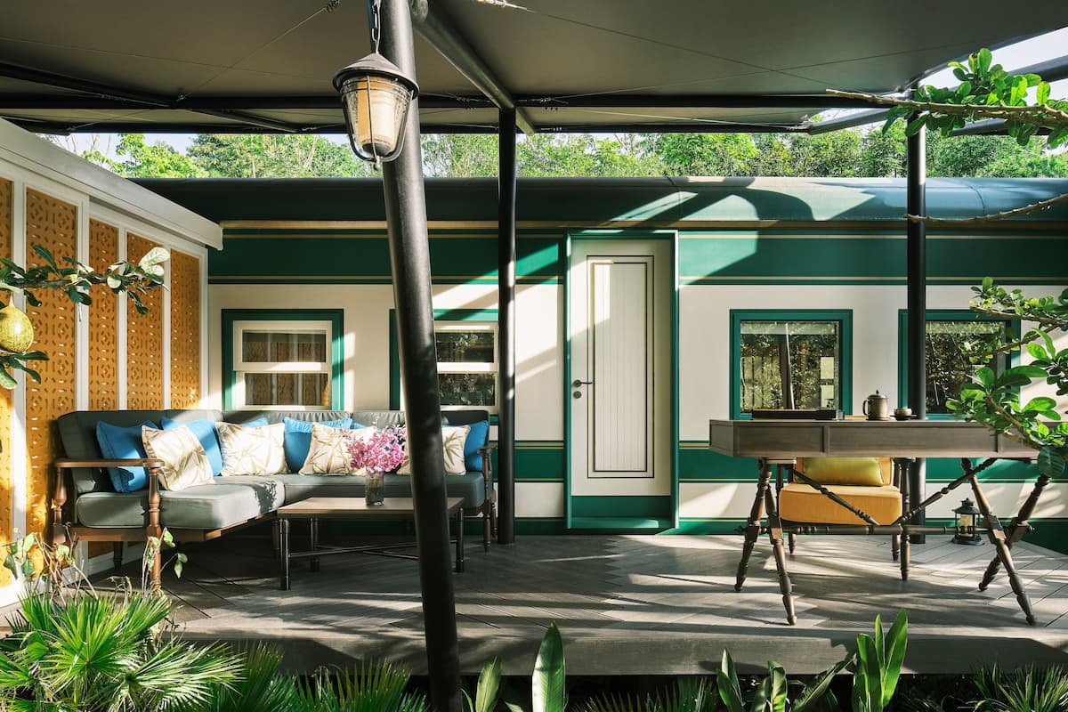 A covered outdoor seating area with a sofa, table, and chairs, surrounded by greenery and a decorative wall.
