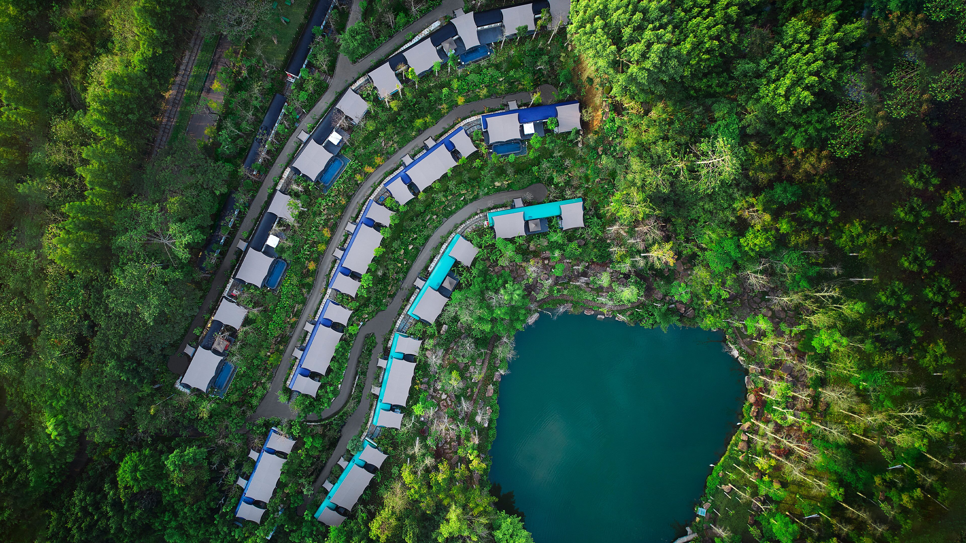 An aerial view of a resort with multiple buildings arranged in a curved layout, a central water body, and lush greenery surrounding the area.